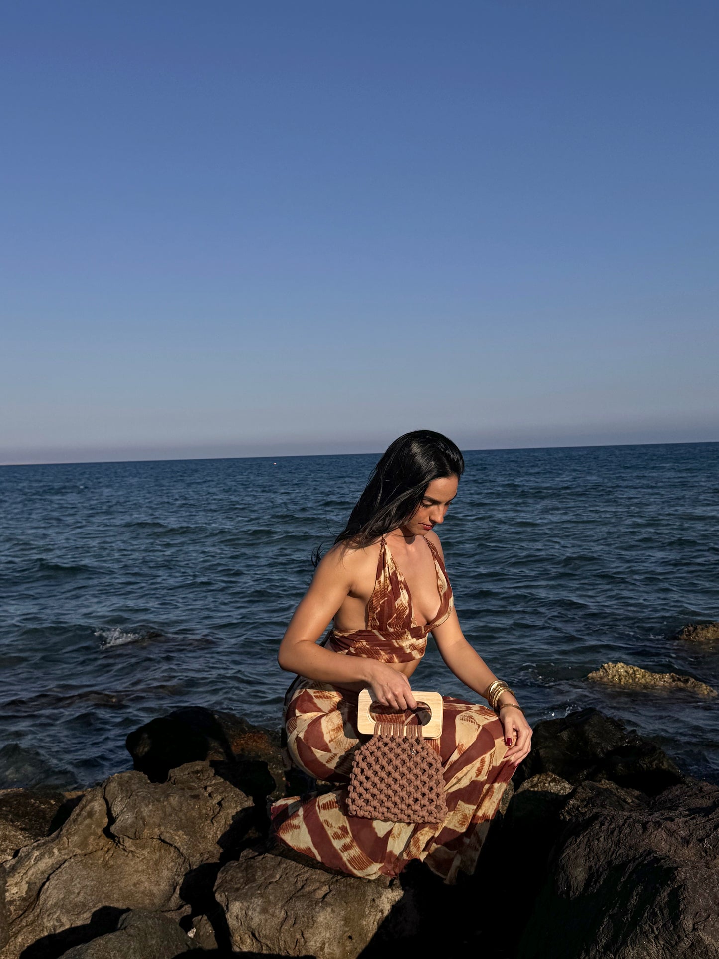Woman sitting on rocks by the ocean holding a macrame bag.