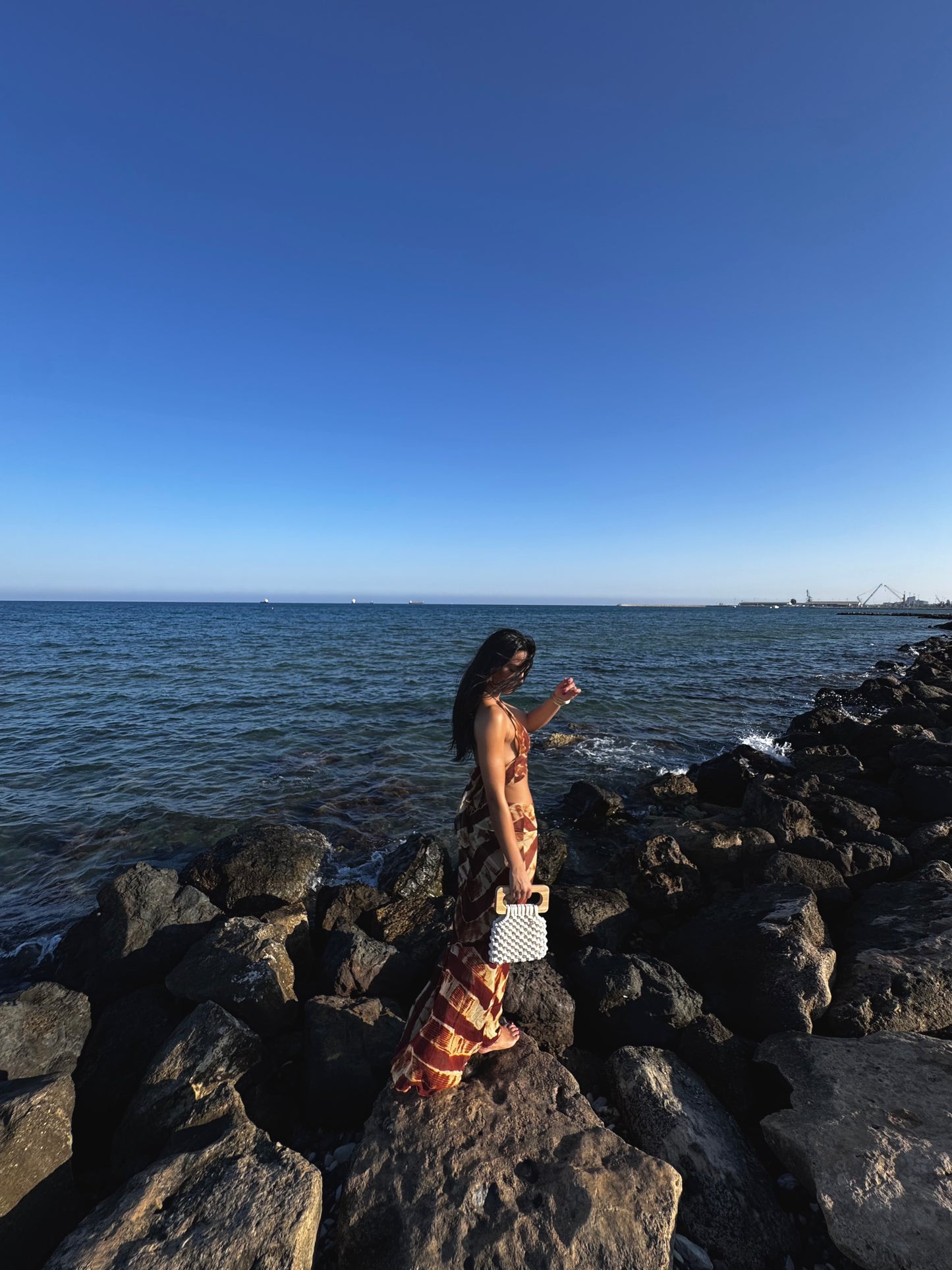 Woman standing on rocks by the ocean with a clear blue sky and her Koma bag