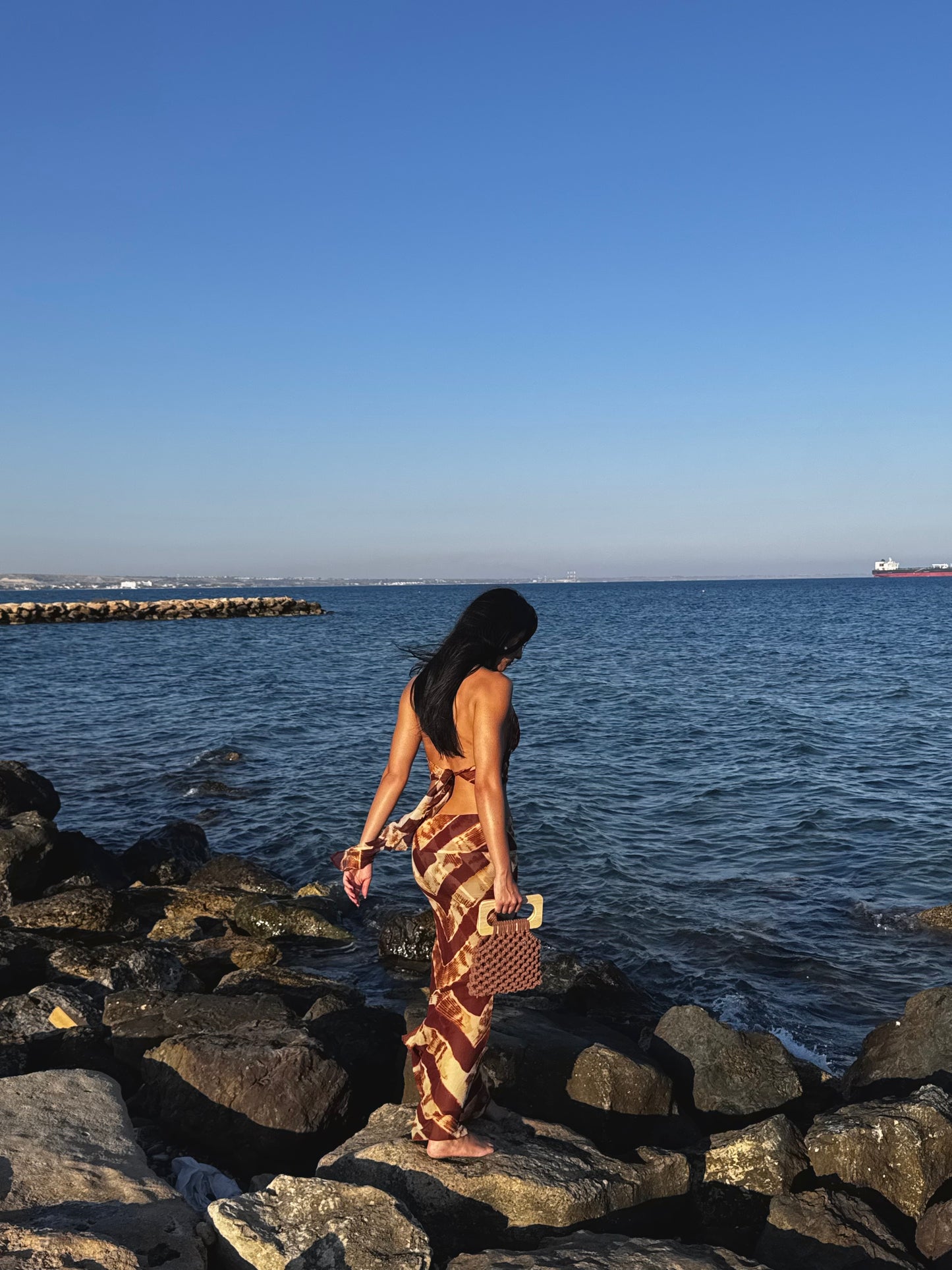 Woman walking along a rocky shoreline with ocean and clear sky in the background WITH THE KOMA BAG