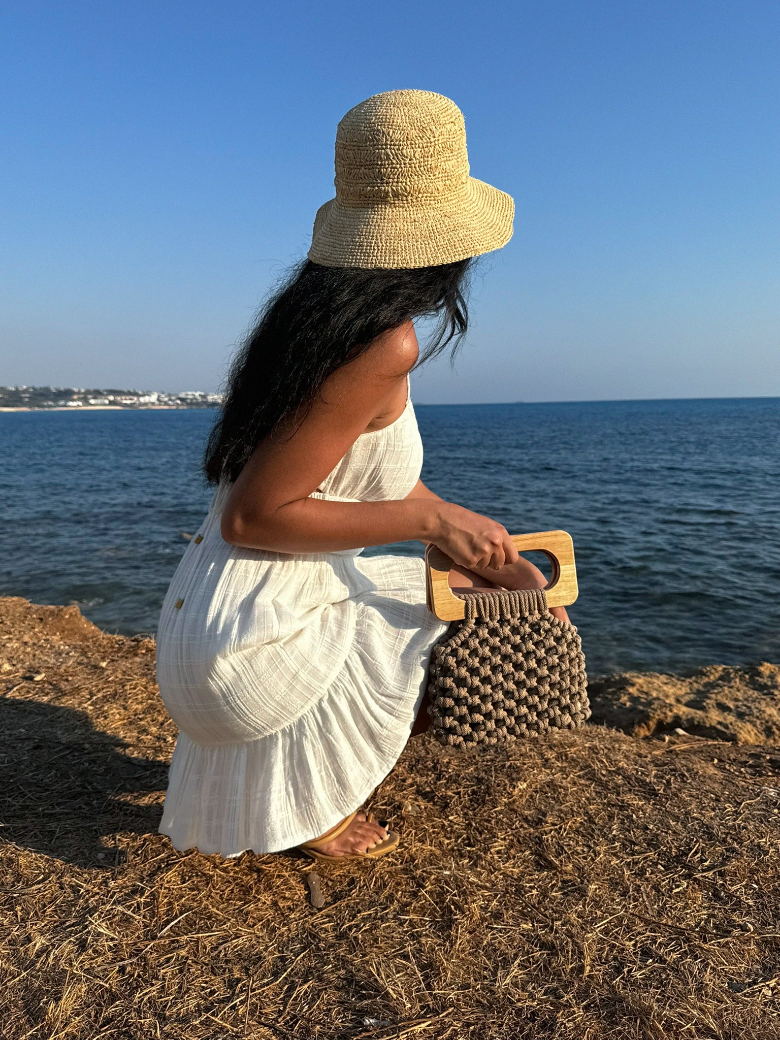Handmade Koma Bag in Sand, styled with a white mini dress and a raffia sun hat and flat sandals. perfect summer vacation outfit.
